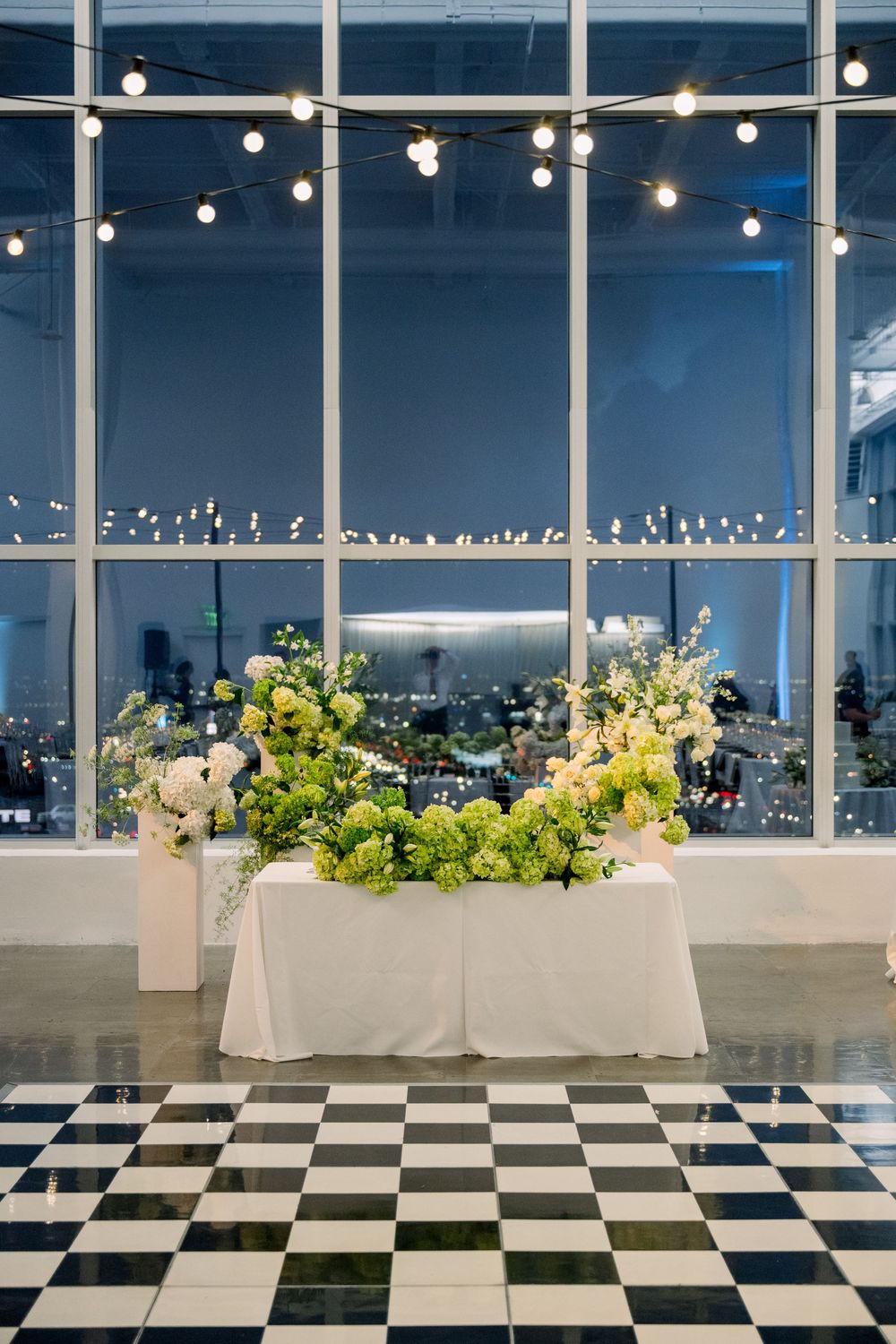 Elegant wedding reception space with black and white checkered floor and floral arrangements under string lights.