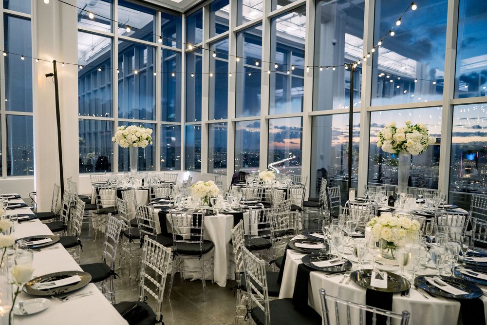 An elegant wedding reception venue with floor-to-ceiling windows, white-clothed round tables, and silver chiavari chairs.