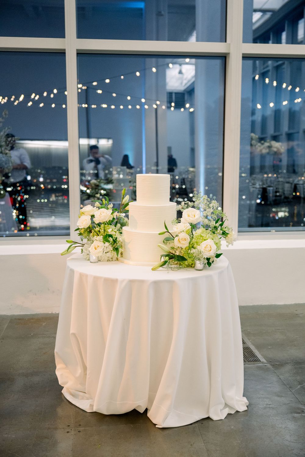 White three-tiered wedding cake displayed on draped table with floral decorations and city lights in background.