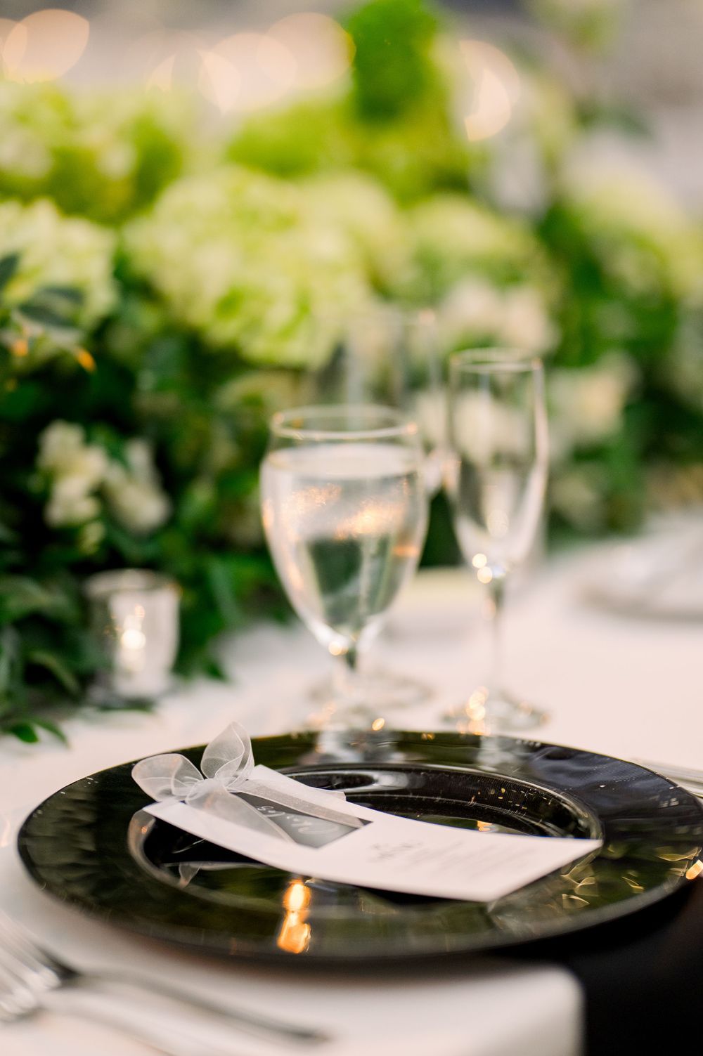 Elegant place setting with black plate, wine glasses and greenery decor at wedding reception.