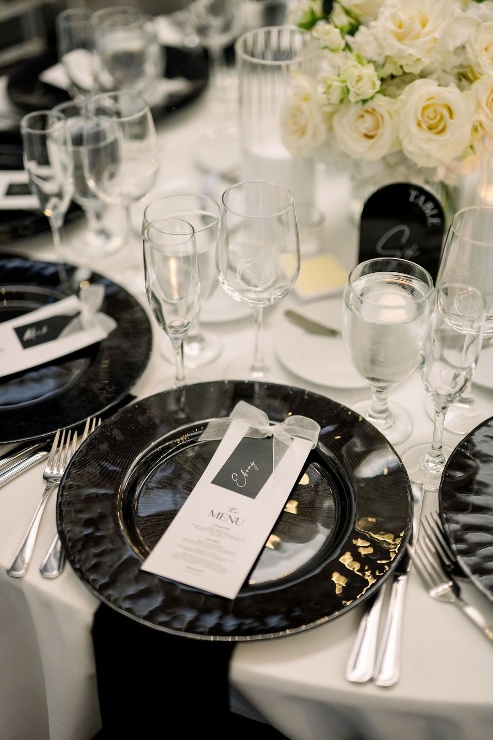 Formal place setting with black charger plate, menu card, and wine glasses on white tablecloth.