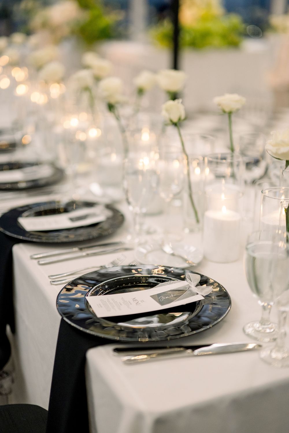 Elegant table setting with black plates, white roses, and candlelight for wedding reception.