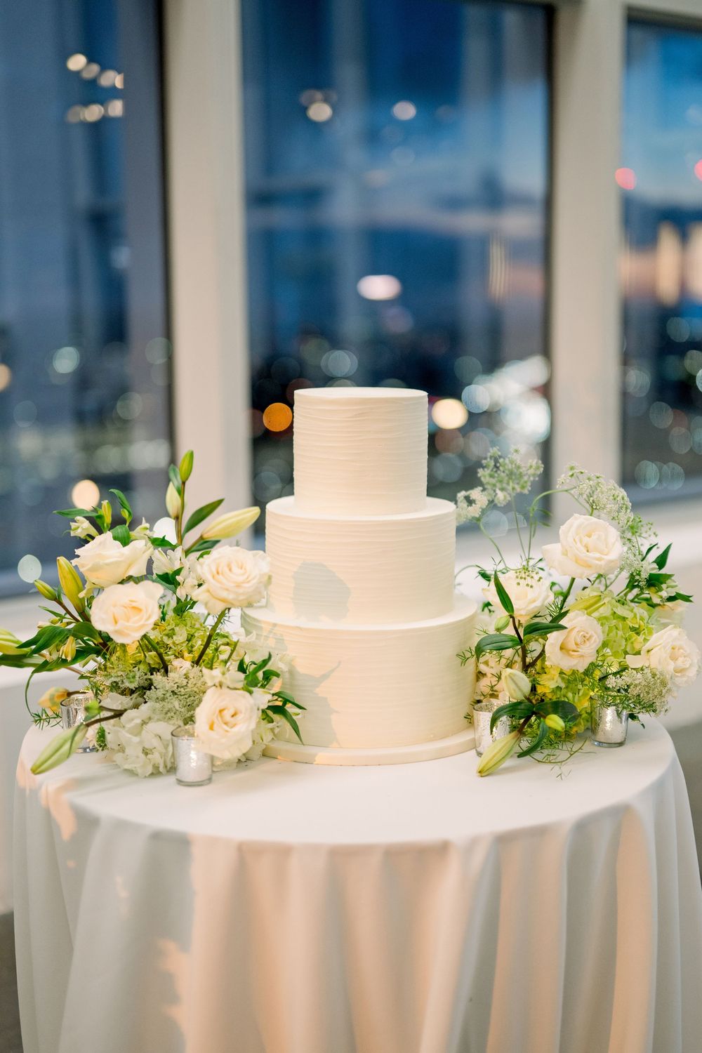 Simple white wedding cake decorated with cream roses and greenery on white tablecloth.