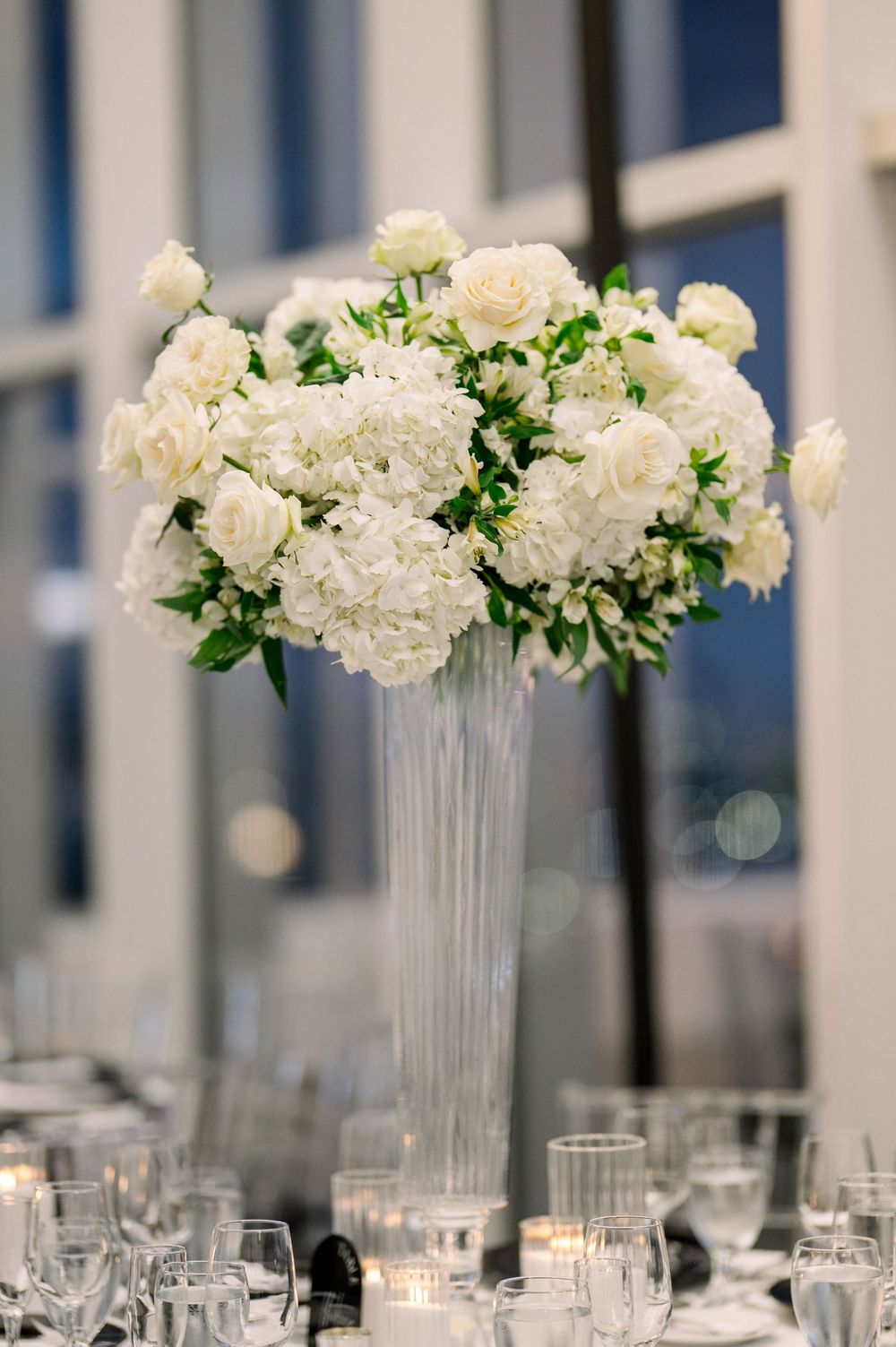 Tall glass vase centerpiece filled with white roses and hydrangeas surrounded by votives.