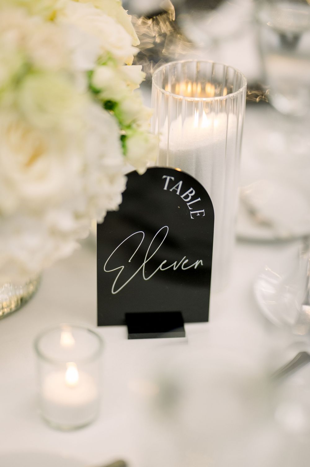 An elegant wedding table setting with a black name card labeled 'Eleven' and glowing votive candles.