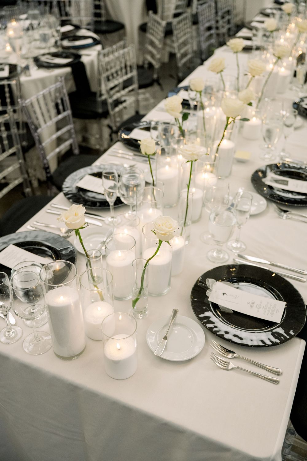 Elegant monochromatic wedding reception table setting with white linens, black plates, and single stem flower centerpieces.