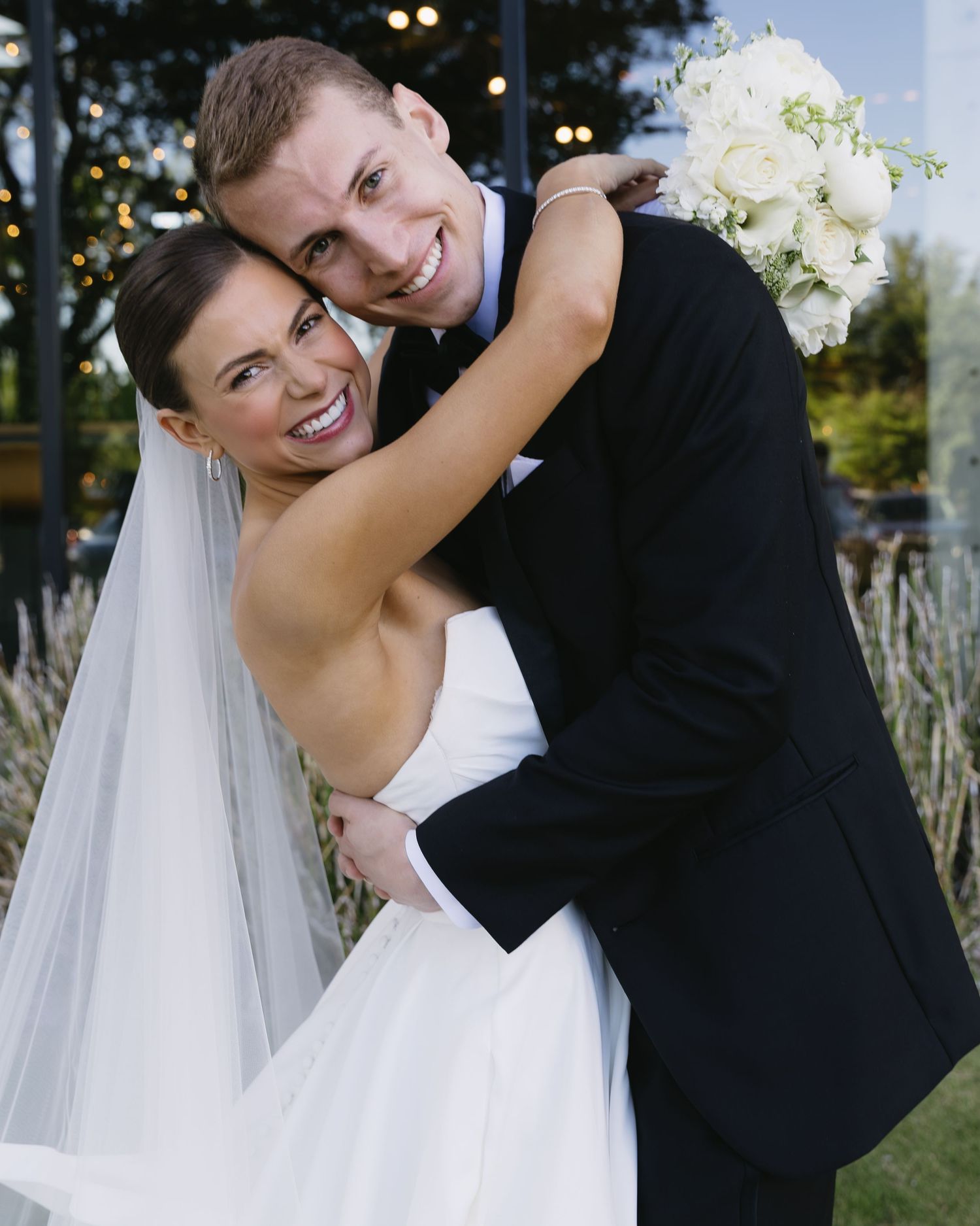 Wedding portrait of smiling bride in white dress with veil and groom in black tuxedo embracing outdoors.
