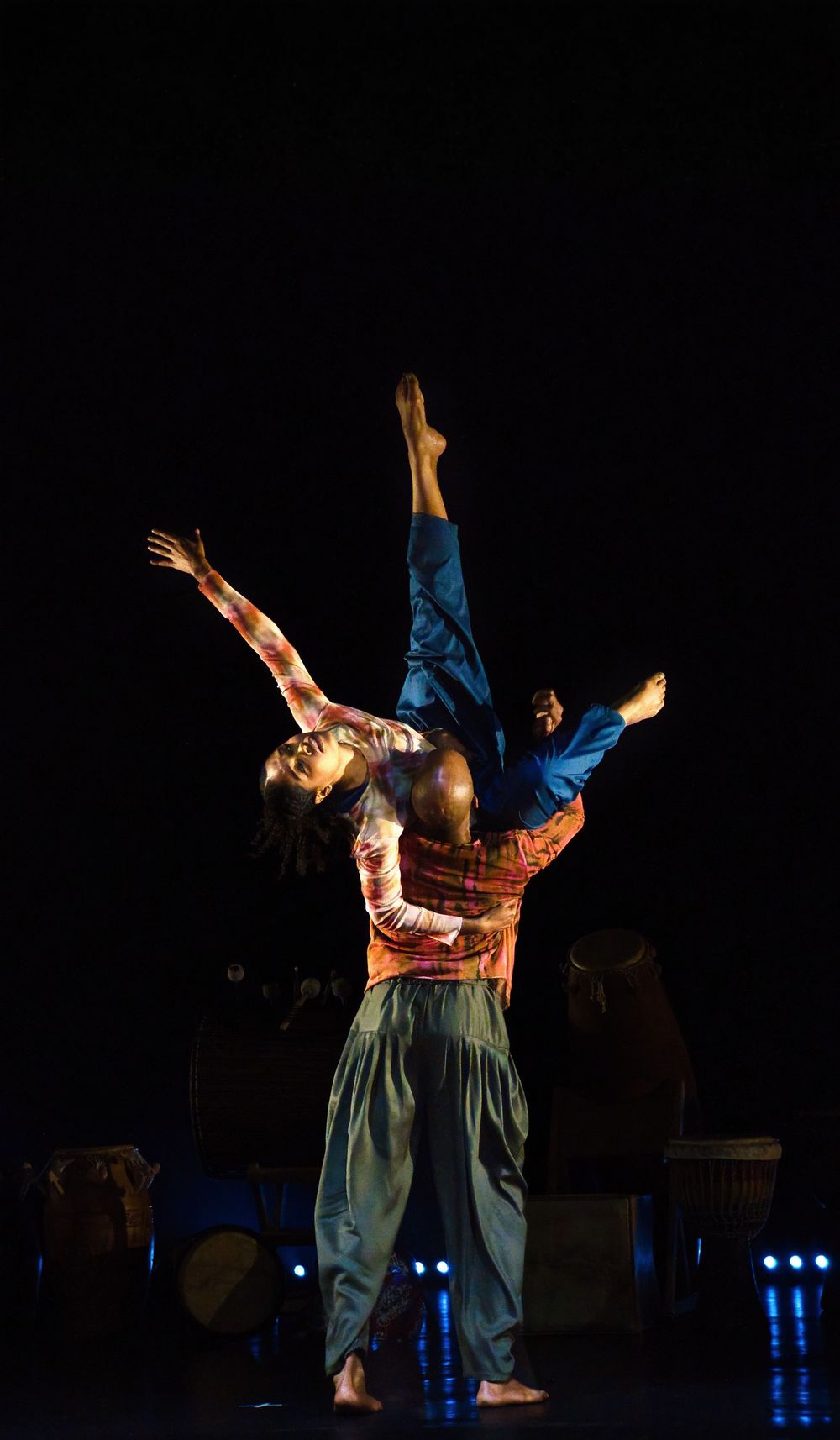 Two dancers perform an acrobatic lift during a dramatic stage performance with blue lighting in the background.