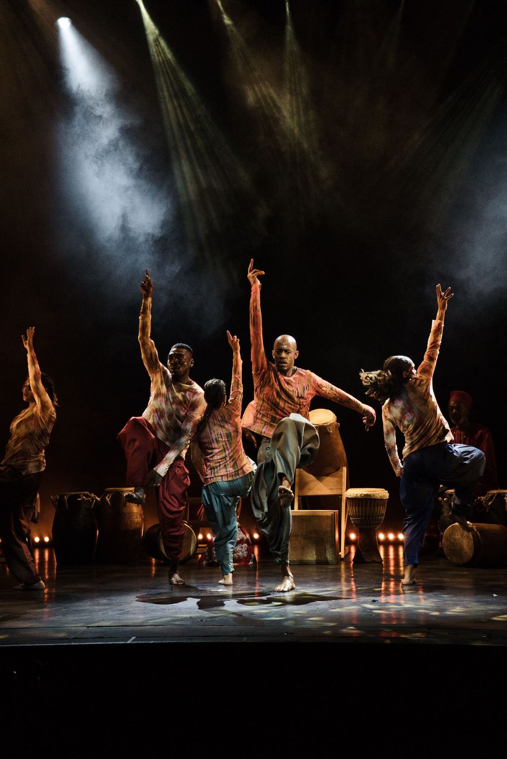 Dancers perform synchronized movements with arms raised under dramatic spotlights on stage.