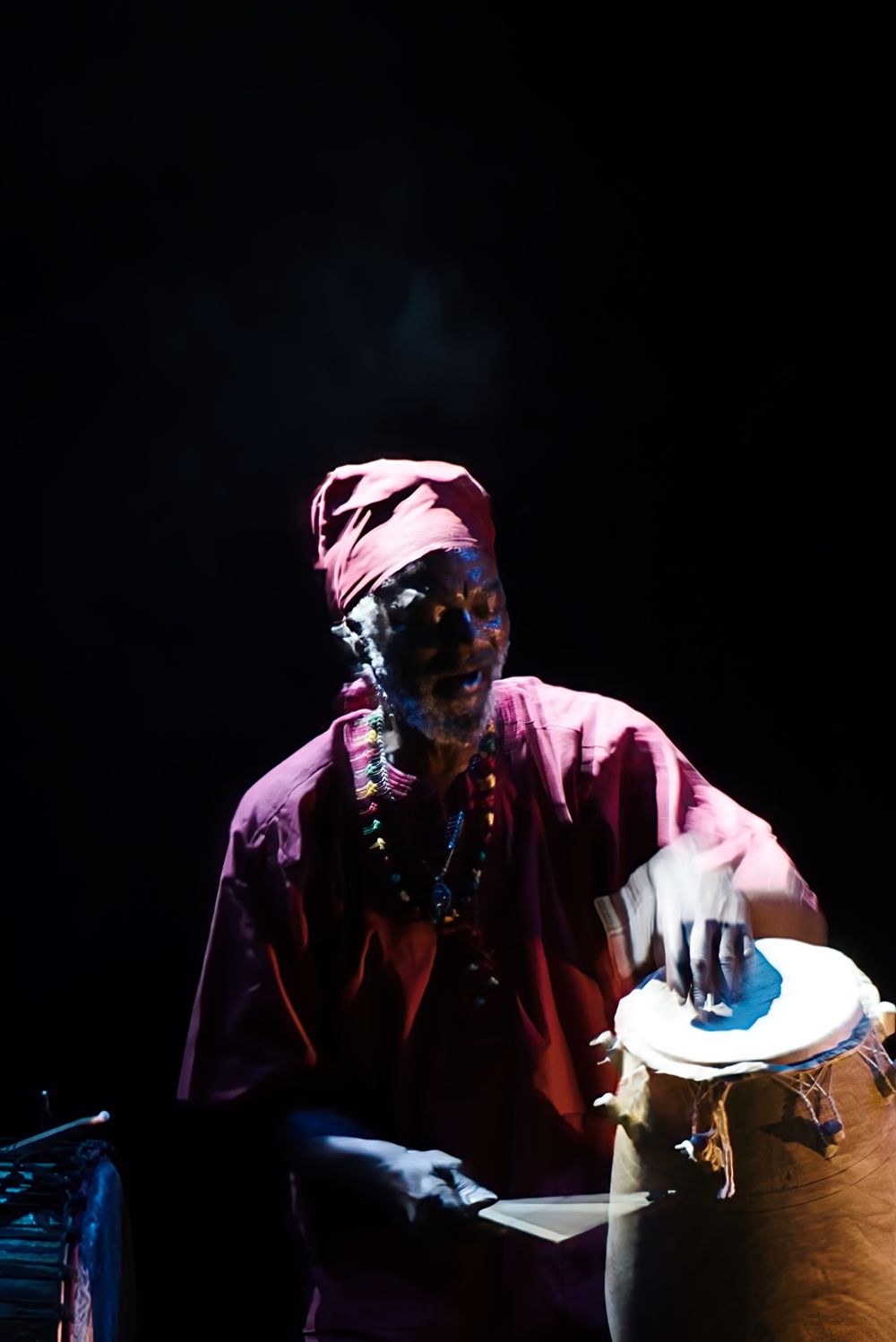 Drummer in traditional dress playing drums under dramatic stage lighting.