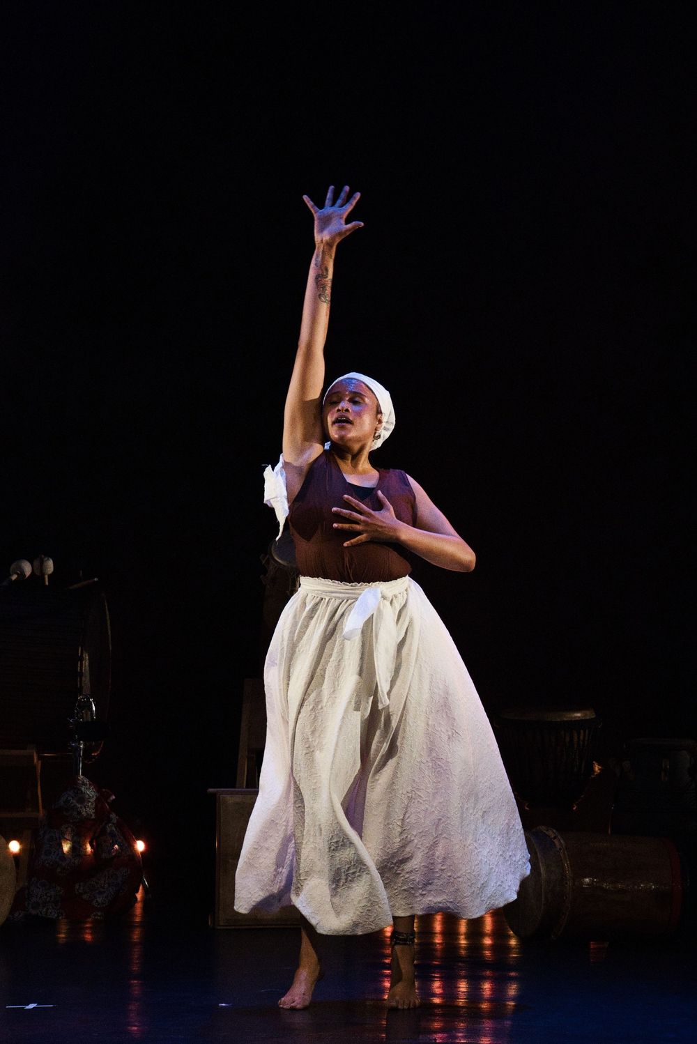 Performer in white flowing dress and headwrap performs expressive dance movement on stage.