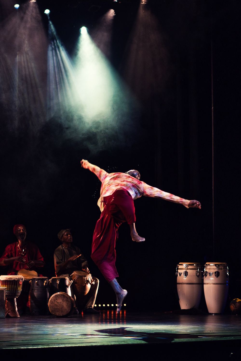 Dynamic jump pose during theatrical performance with percussion instruments visible in background.