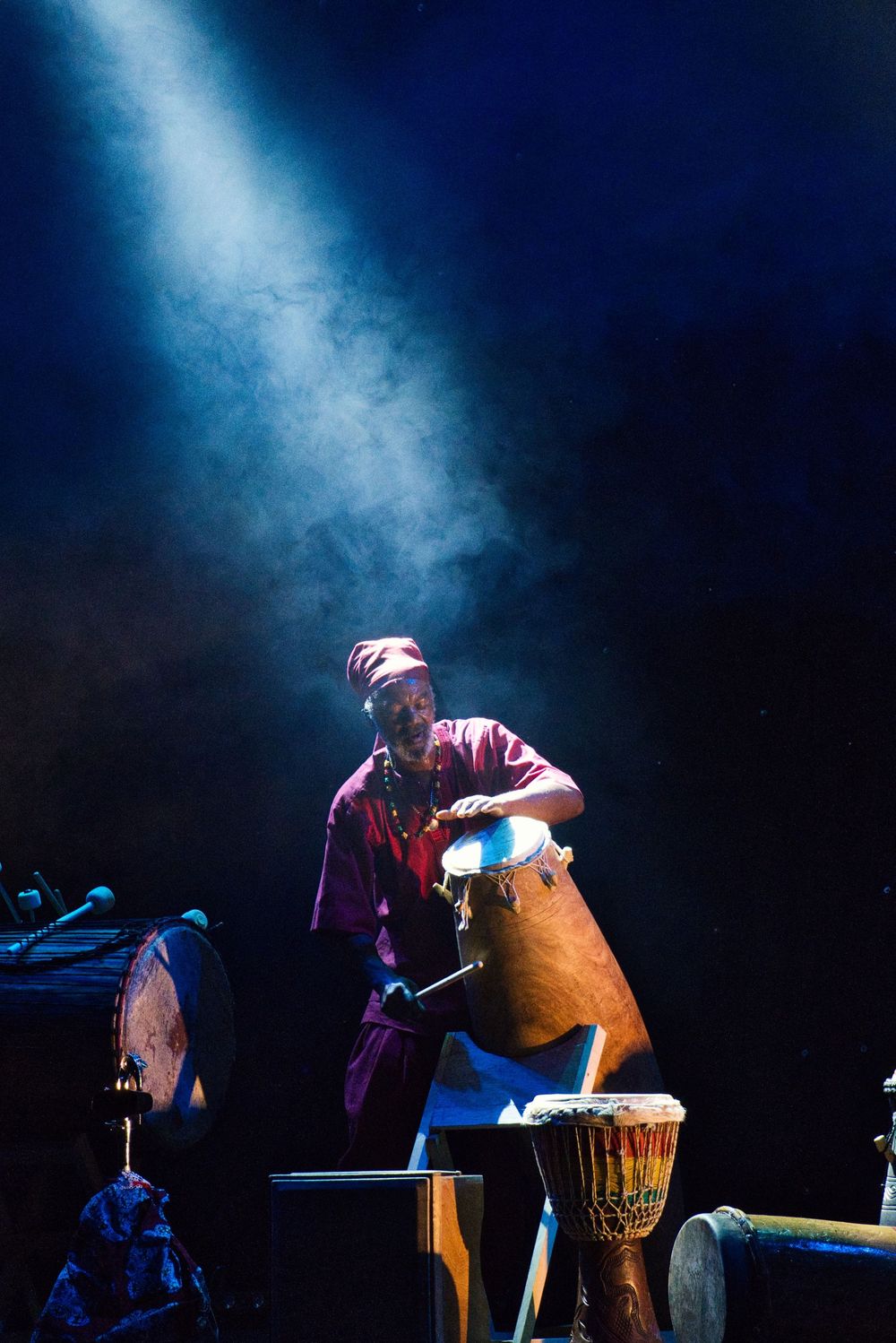Percussionist playing traditional drums under blue spotlight on dark stage.