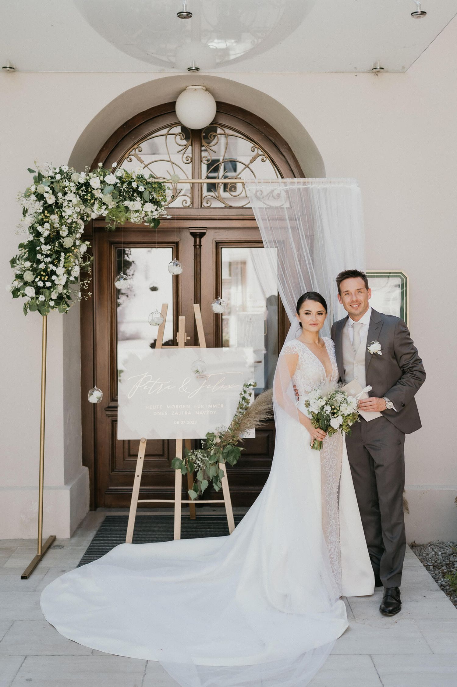 Elegant newlywed couple poses in front of arched doorway with white floral decorations and seating chart display.