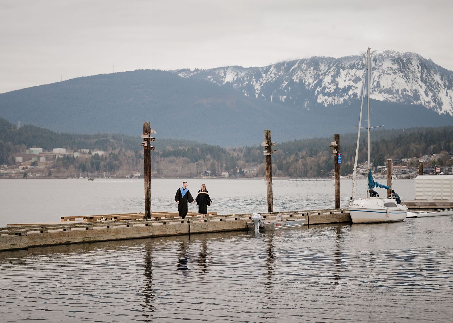 Two people on a wooden dock overlooking a scenic mountain lake with snow-capped peaks in the background.