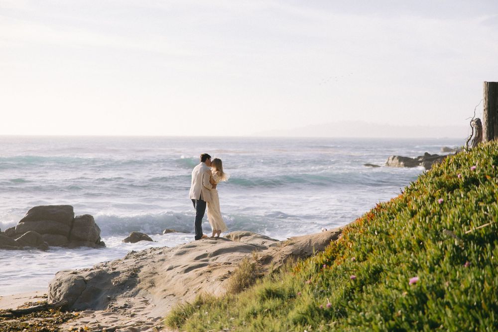 A couple stands on rocky coastal cliffs overlooking the Pacific Ocean as waves crash against the shore on a misty day.