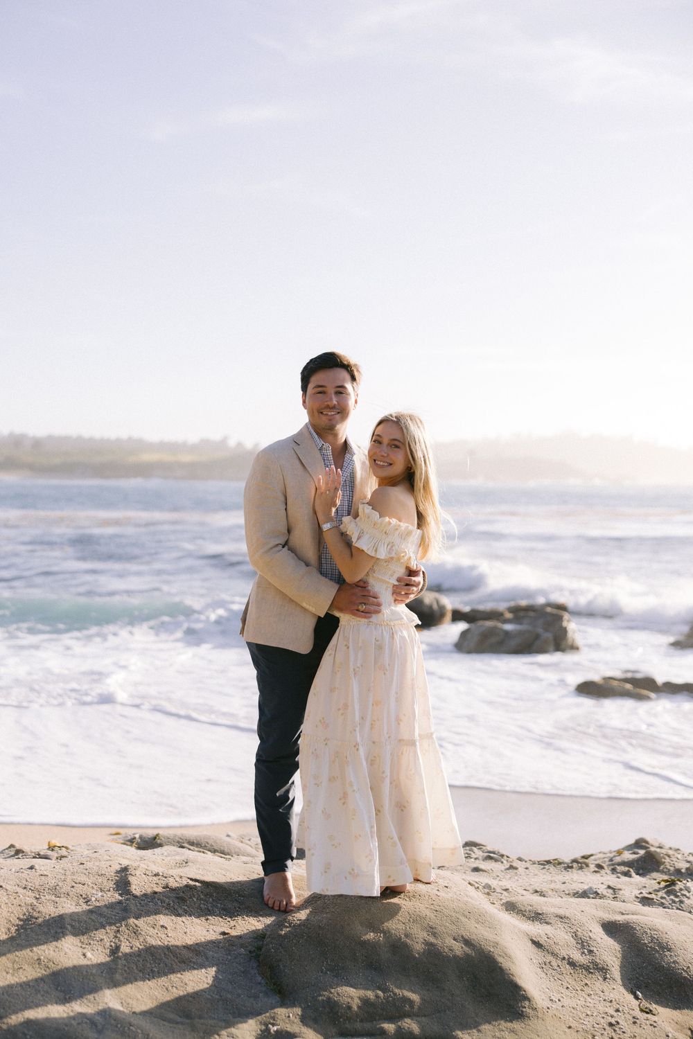 A couple shares a romantic moment together on a rocky beach during sunset with crashing waves in the background.