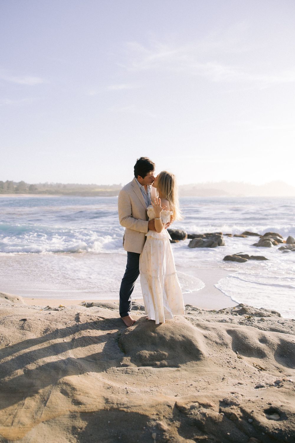 A couple shares an intimate moment while standing on coastal rocks at sunset by the ocean waves.