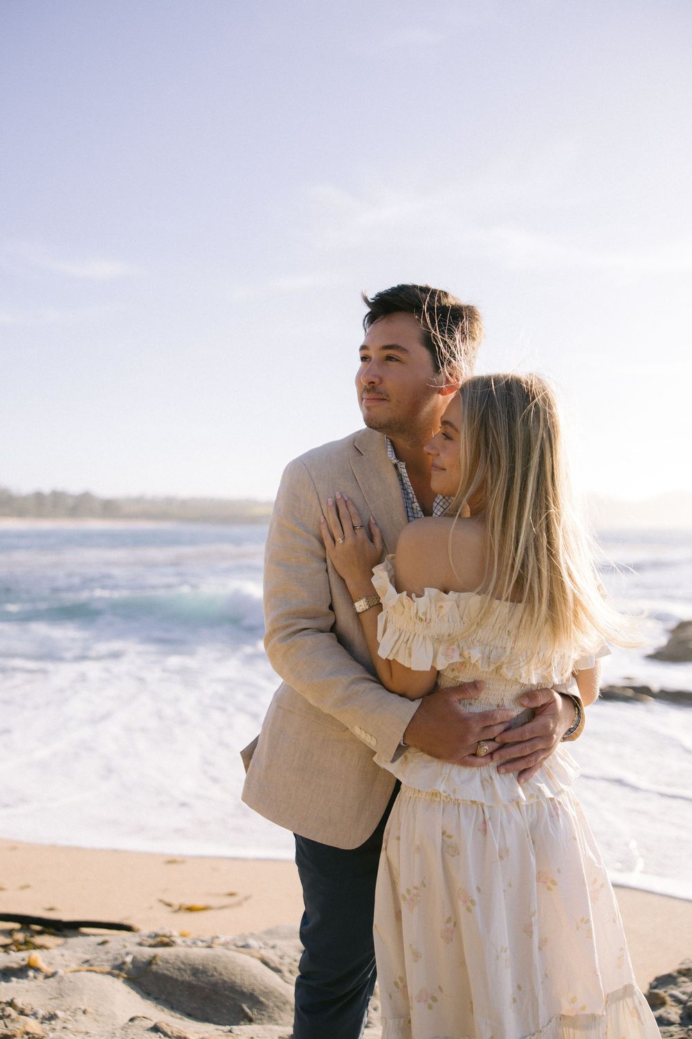A romantic couple embraces on a scenic beach with crashing waves and cloudy skies in the background.