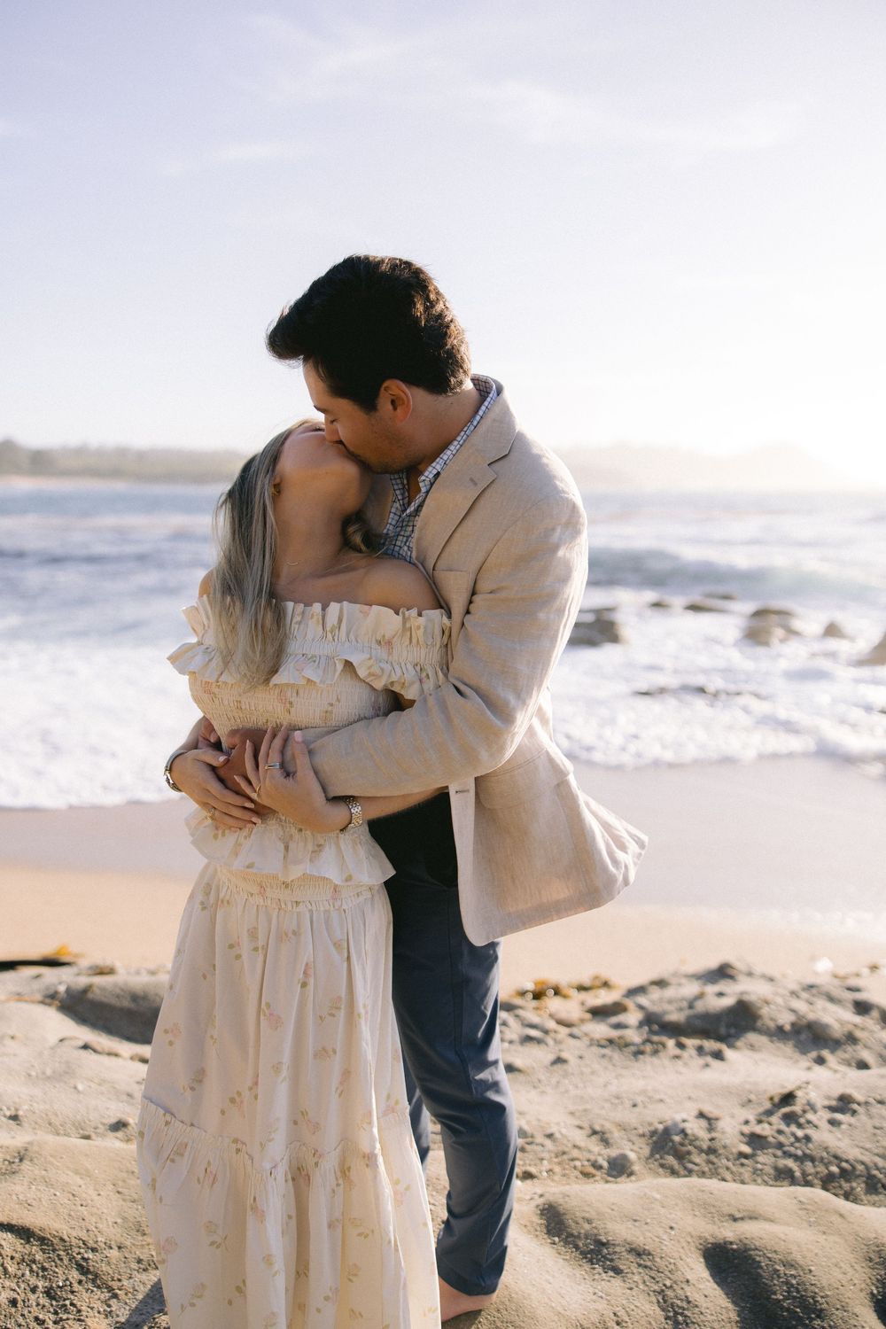 A romantic couple embraces on a beach at sunset, with the woman wearing a flowy white dress and man in a beige blazer.