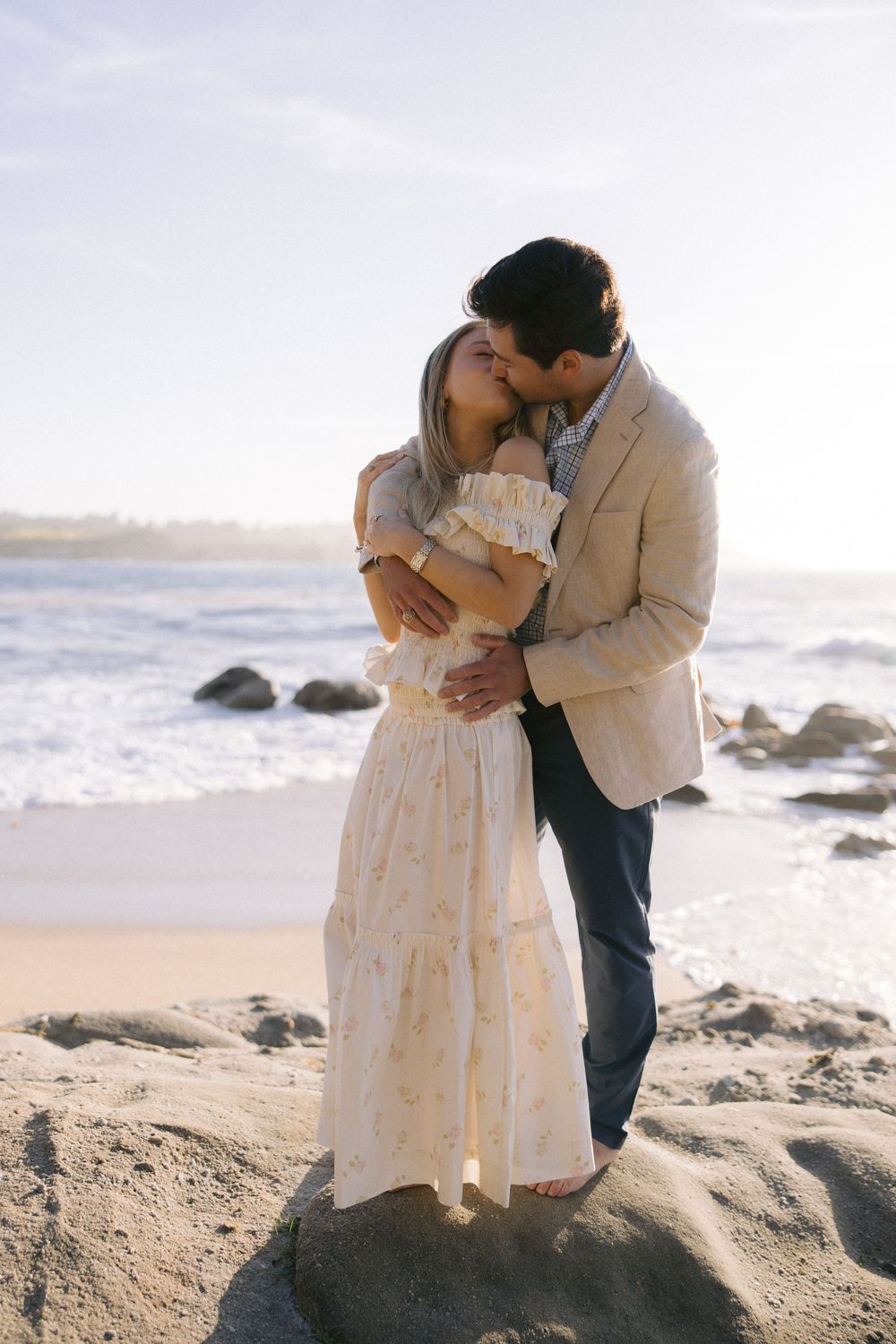 A romantic couple in wedding attire embrace on a rocky beach at sunset with ocean waves in the background.