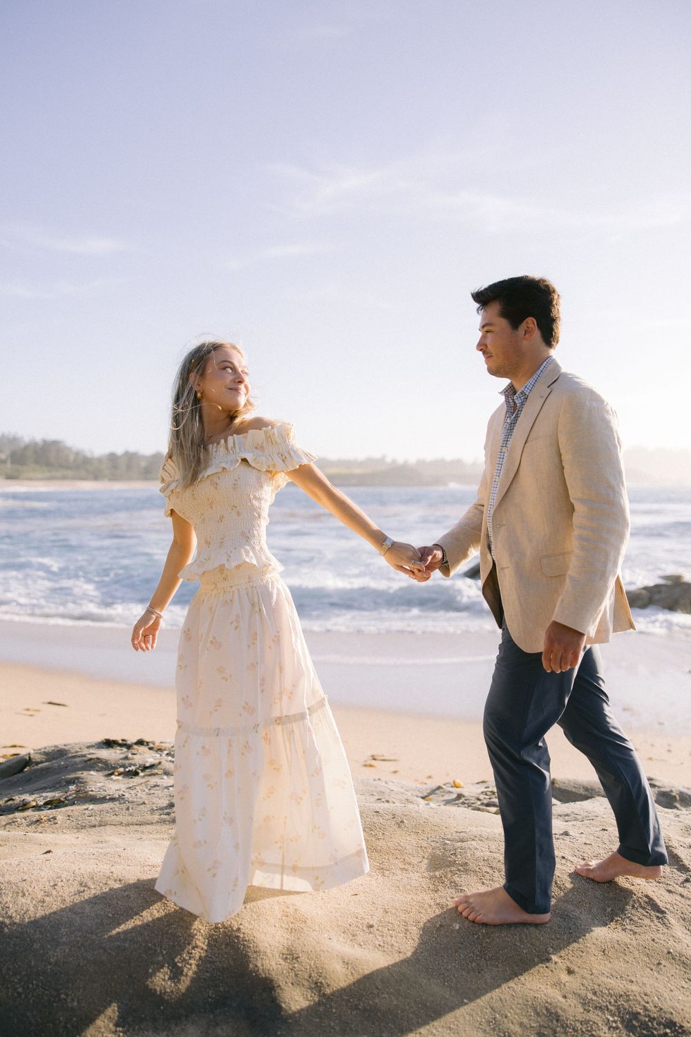 A couple dancing on a sunny beach with waves and rocks in background, wearing vintage-inspired formal attire.