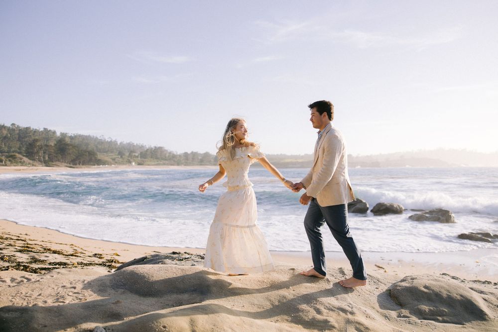 A couple holds hands while walking together on a rocky beach at sunset with crashing waves in the background.