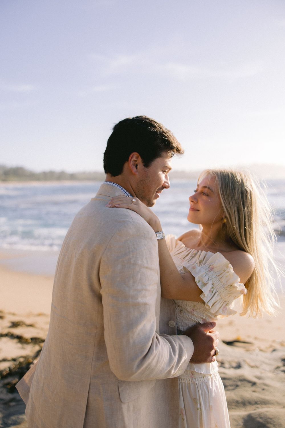 Romantic couple embraces on sandy beach at sunset wearing beige and white outfits.