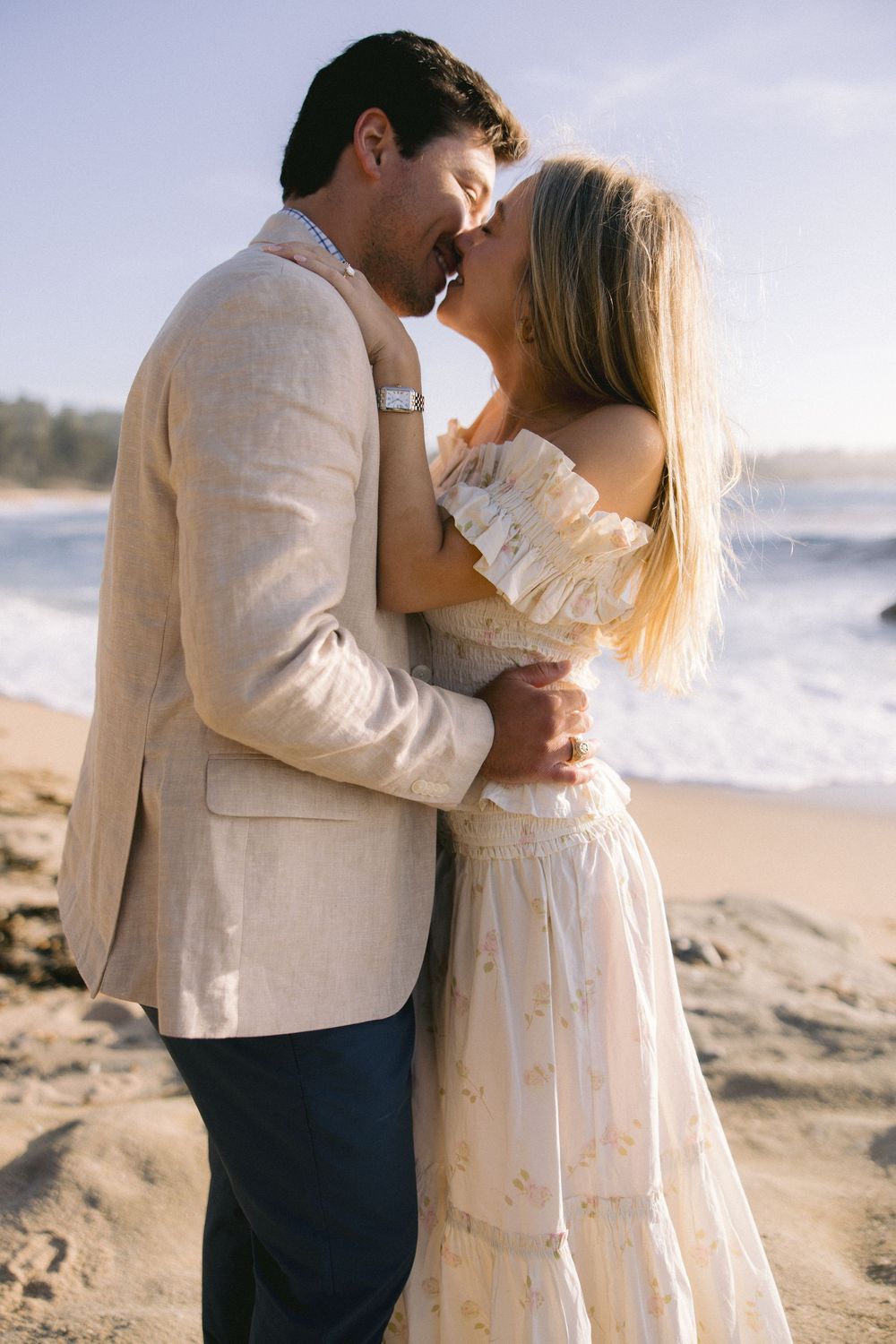 A romantic seaside embrace between a couple in light colored formal wear on a sandy beach at sunset.