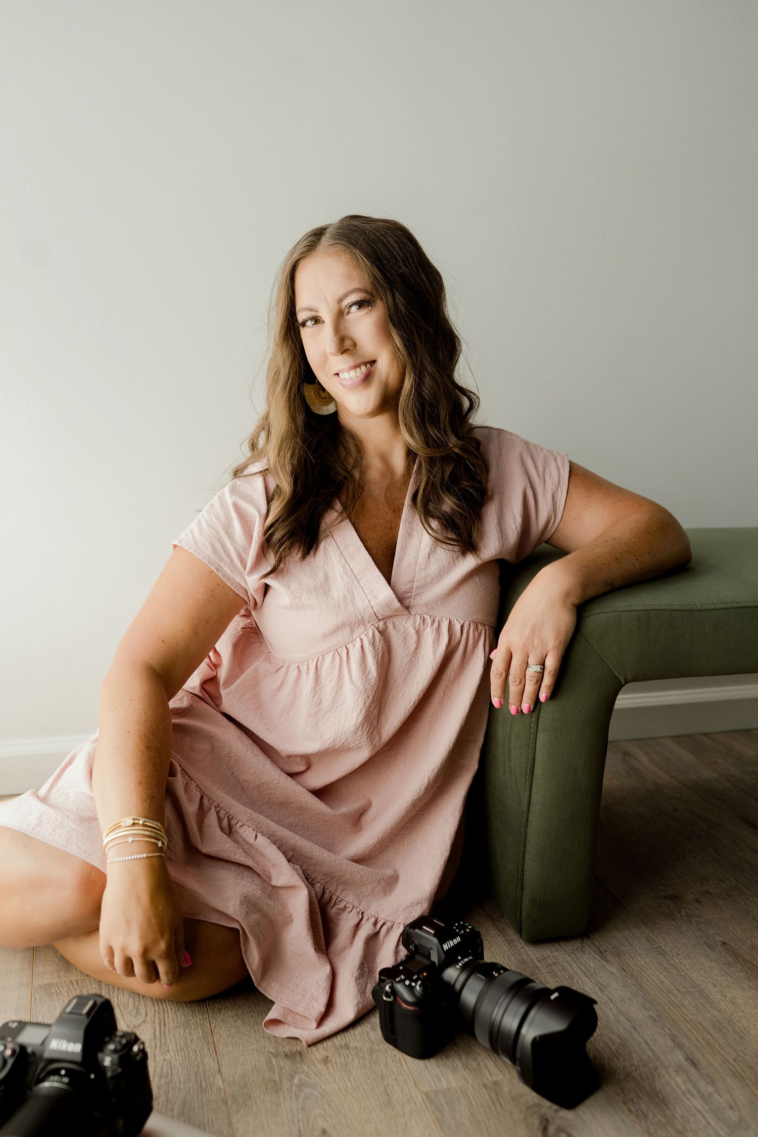 Photographer in pink dress poses casually with camera on grey couch.