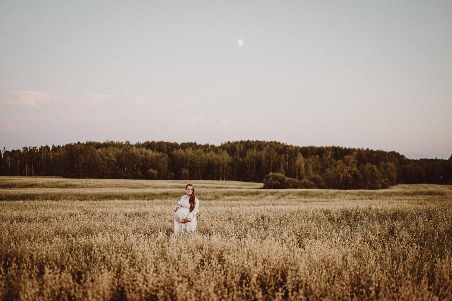 Photo de maternité d’une femme en blanc debout dans un champ doré au crépuscule.