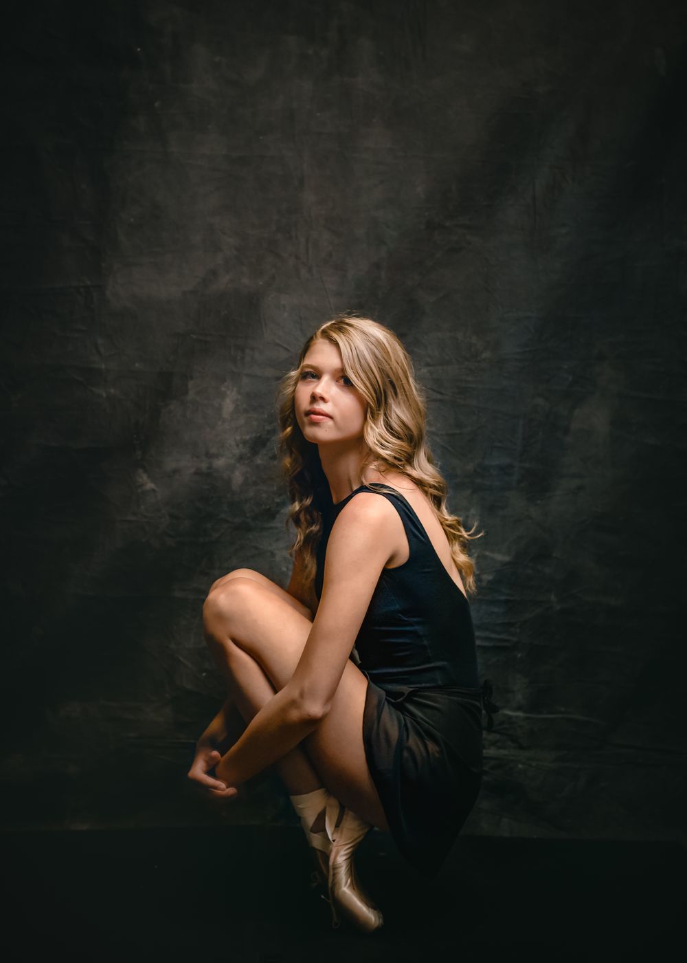 Artistic dance portrait featuring a ballerina in a black dress crouching gracefully against a dark textured background.