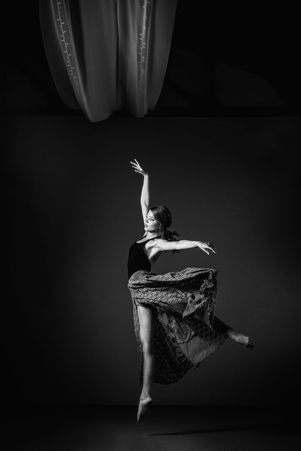 A black and white artistic photograph of a dancer performing a dynamic leap pose in a flowing dress against a dark background.