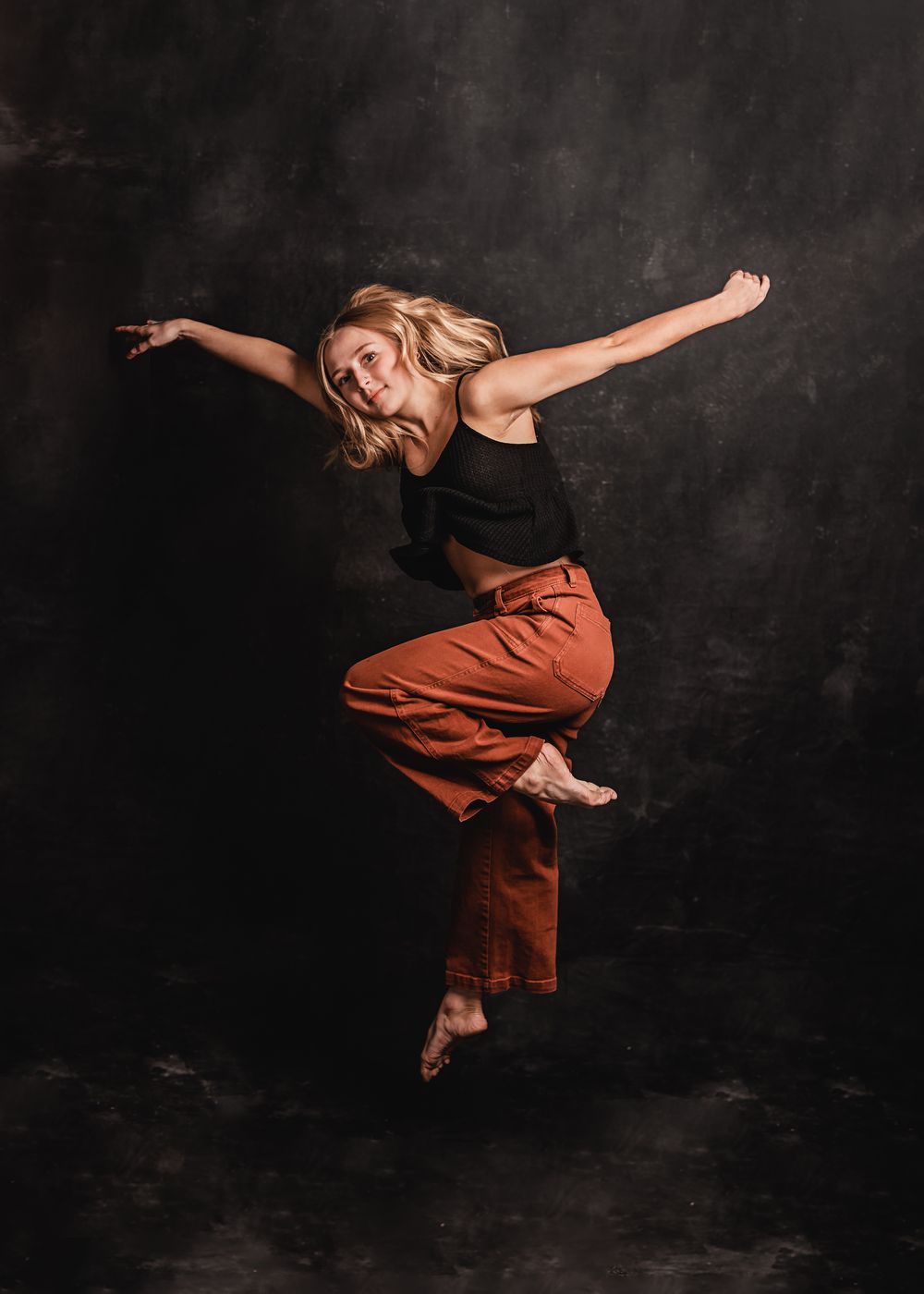 Dancer in black top and rust-colored pants leaps gracefully against dark backdrop in dynamic contemporary dance pose.