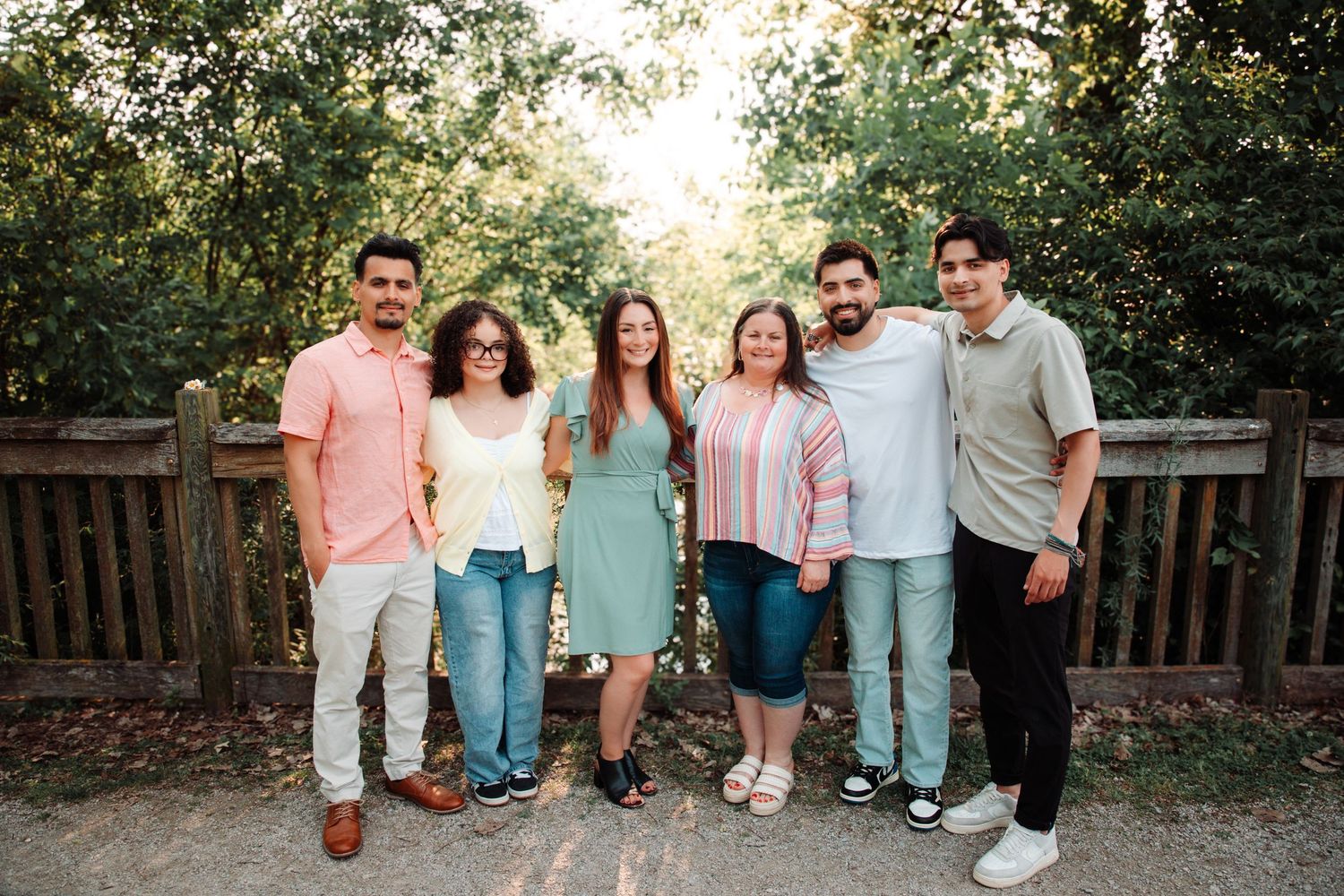 Group portrait of friends in casual summer attire standing on wooden deck with sunlit trees behind.