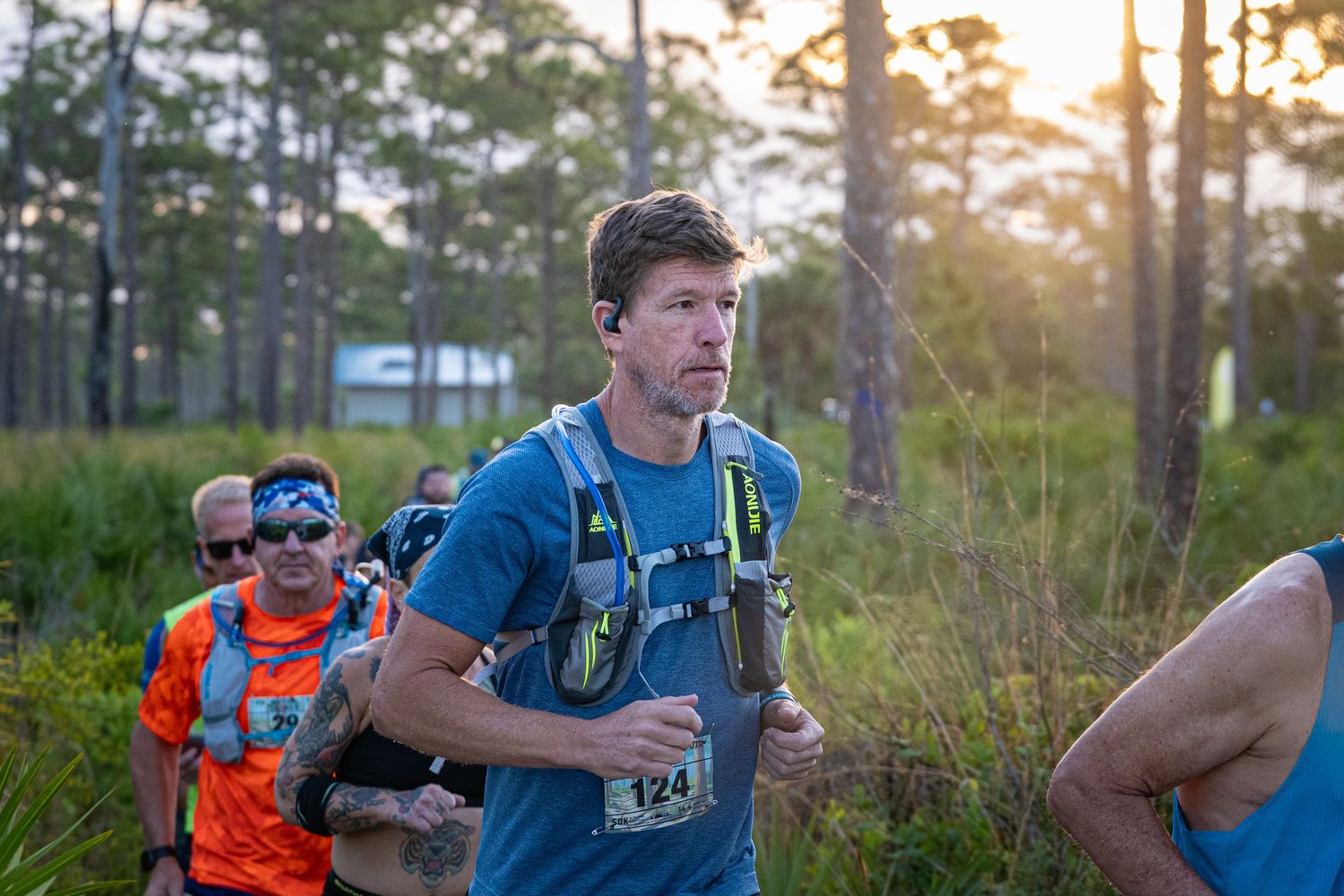 Trail runners competing in an endurance race through pine forest at sunset wearing hydration packs and race bibs.