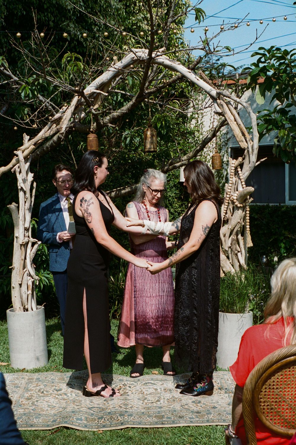 Wedding ceremony taking place under a natural wood arch with hanging lanterns in a garden setting.