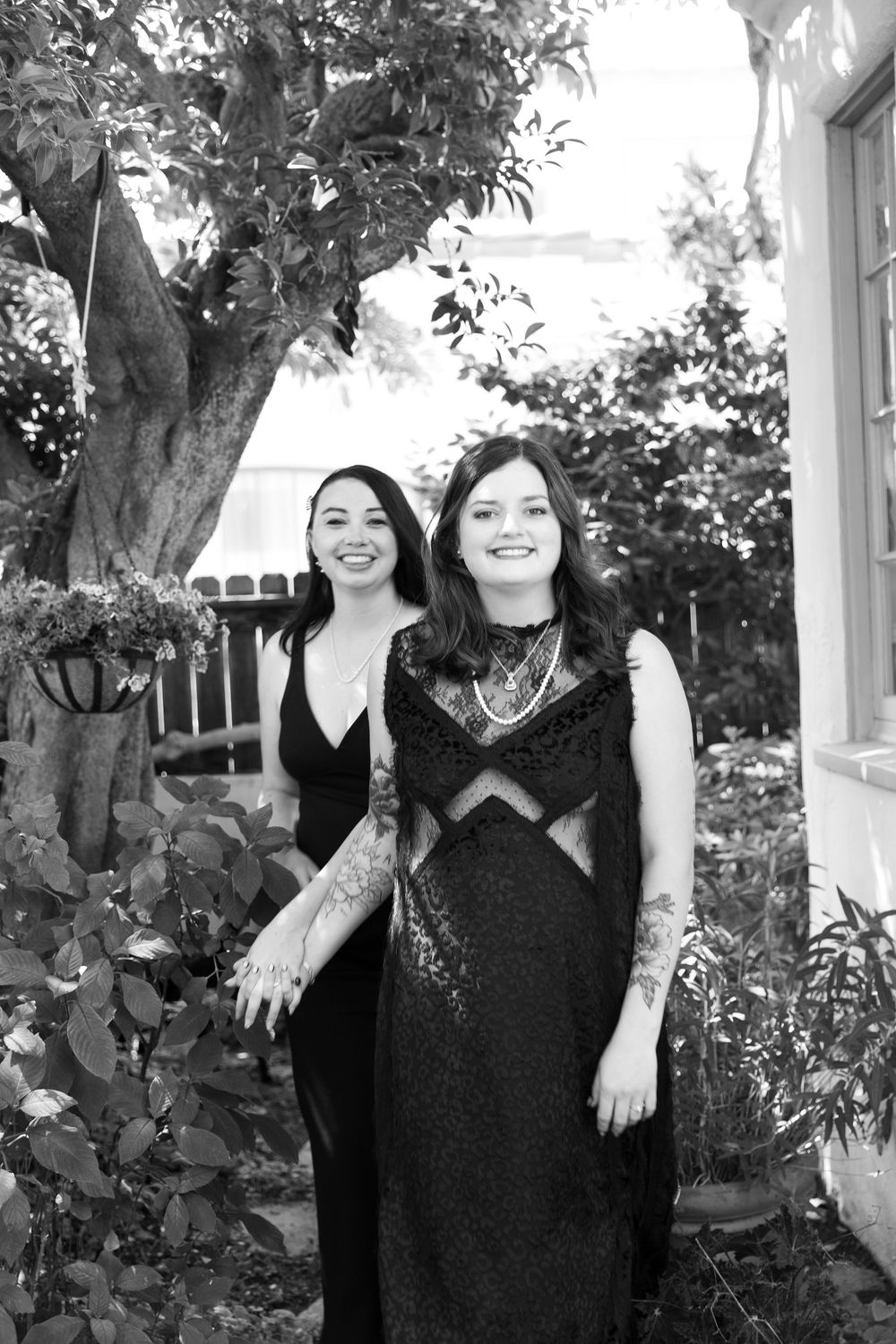 Two people pose together outdoors in front of lush greenery.