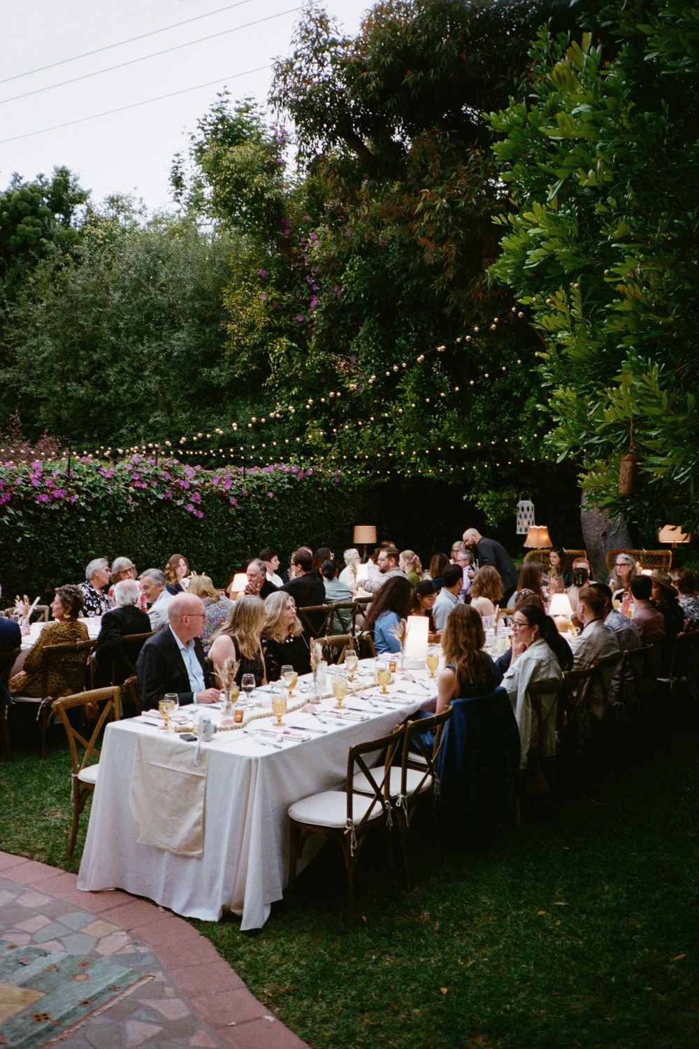 Outdoor dinner party with long white tables and string lights in a garden setting.