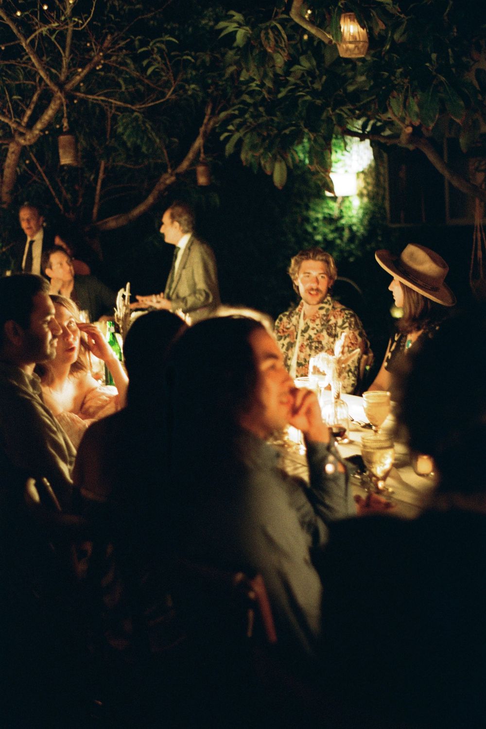 A candlelit outdoor dinner gathering with guests seated around tables beneath garden trees.