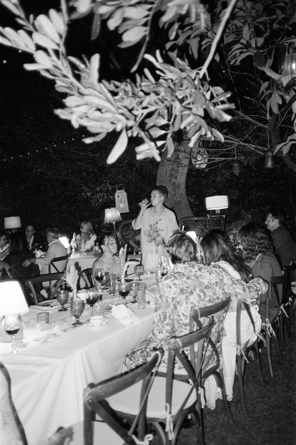 Black and white photograph of an outdoor dinner party with tables set under hanging lights.
