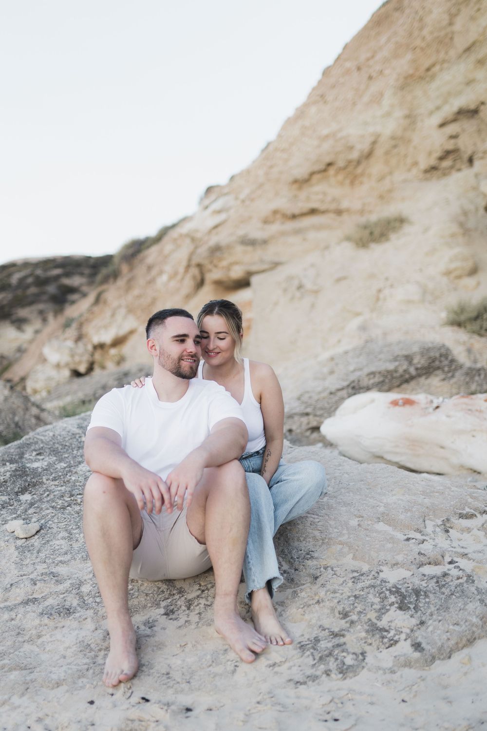 A couple sits intimately on rocky cliffs with a white stone formation in the background during a photoshoot.