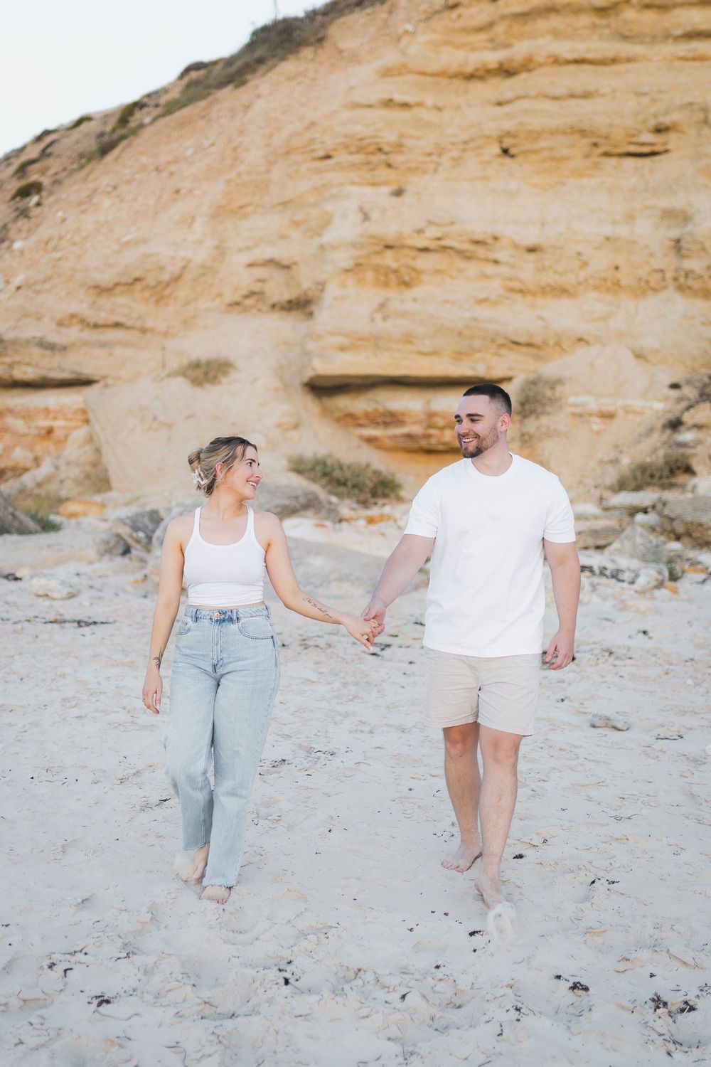A couple walks hand in hand on a sandy beach with rugged sandstone cliffs in the background on a sunny day.