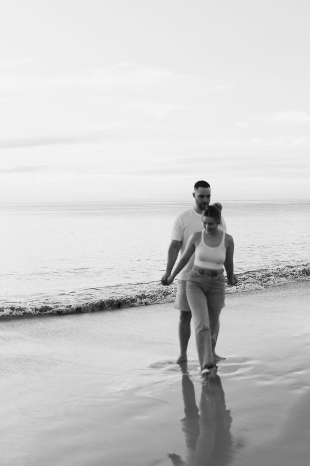A couple takes a romantic stroll along the shoreline at sunset, their reflections visible in the shallow water.