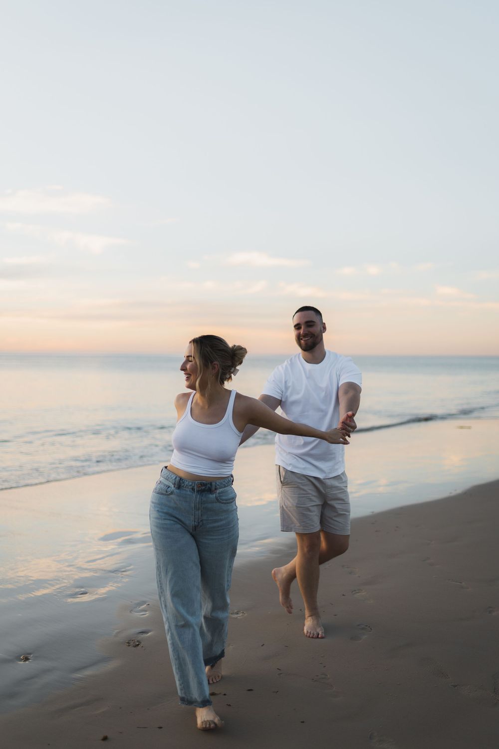 A couple runs playfully along the beach at sunset wearing casual outfits of white tops and denim.