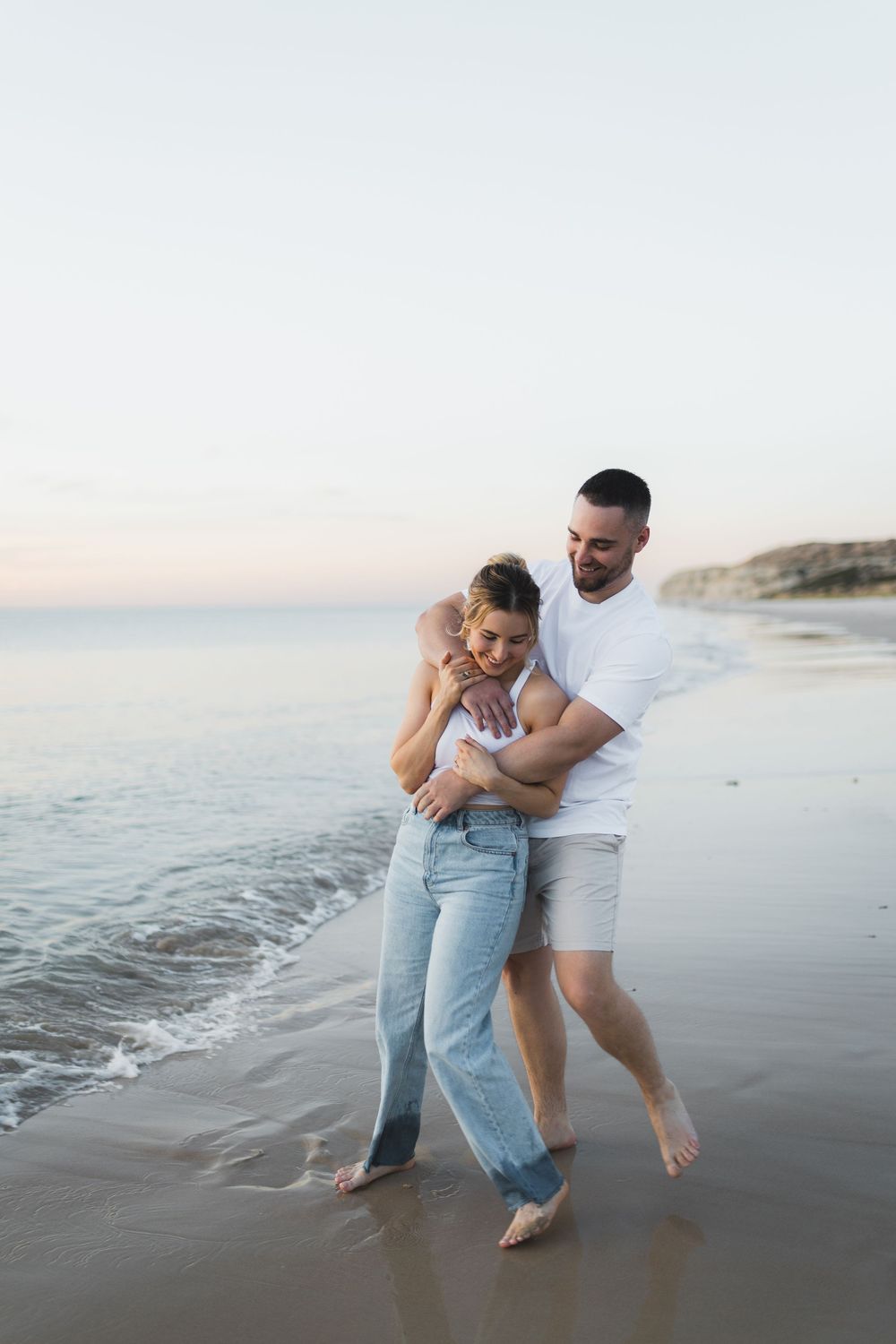A couple embraces and laughs together on a peaceful beach shoreline at dusk.