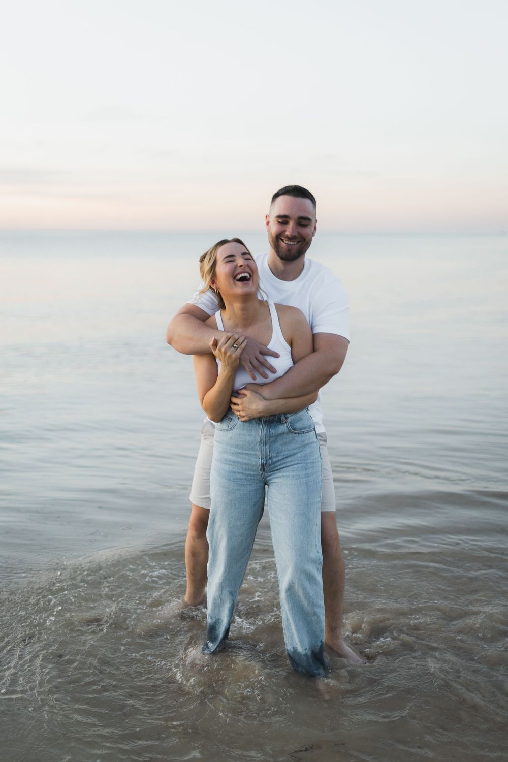 A couple shares a joyful embrace while standing in shallow ocean water at sunset.