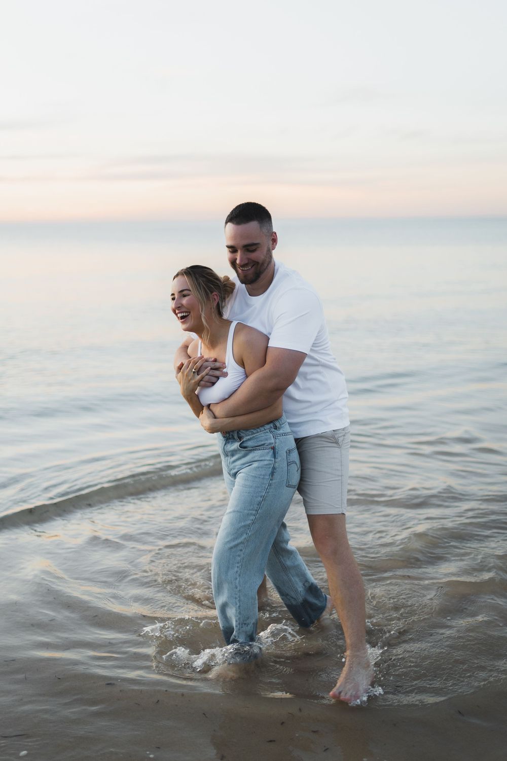 A couple embraces and laughs while standing in shallow ocean waters at sunset.