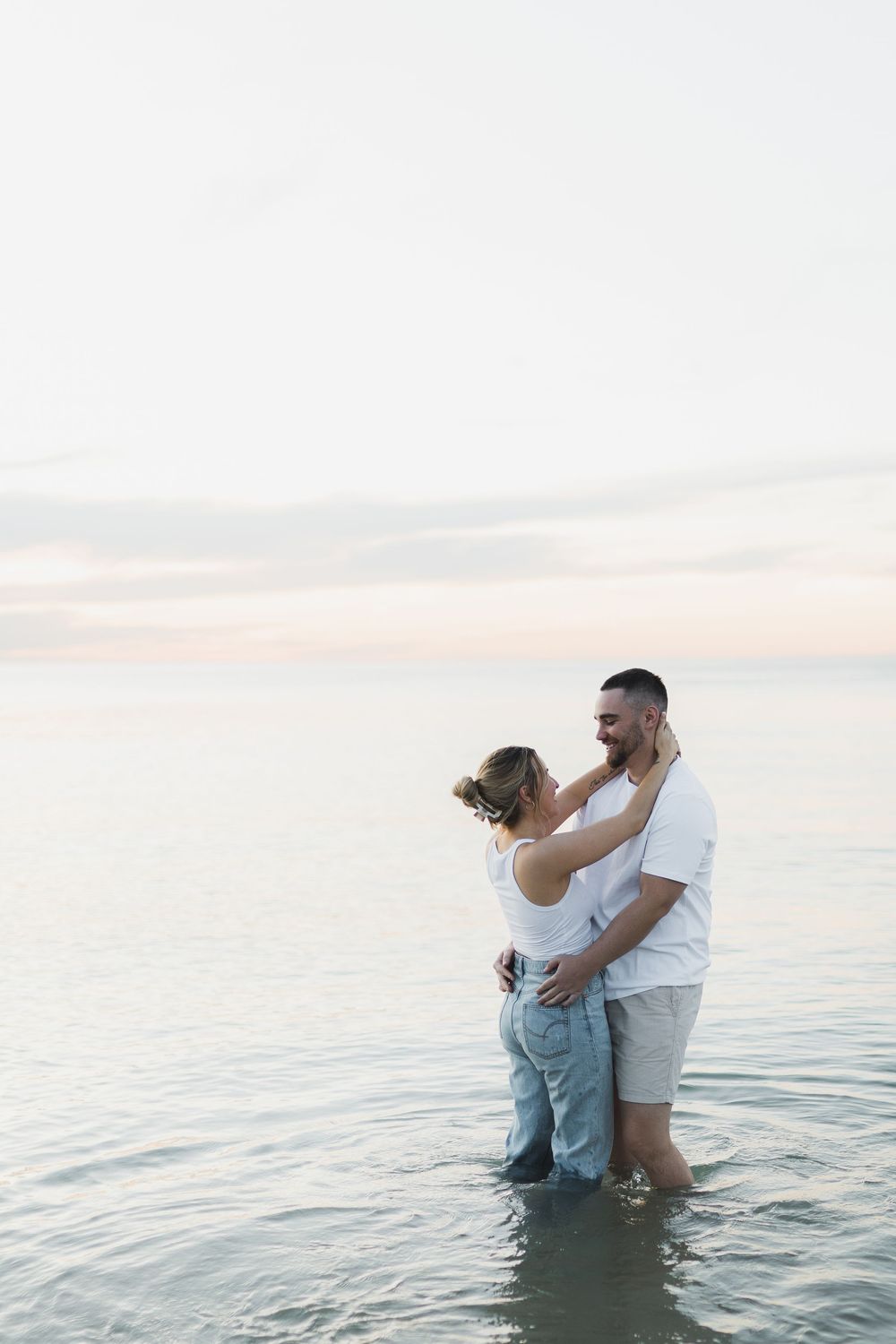 Two people embracing while standing in shallow ocean water during a serene sunset.