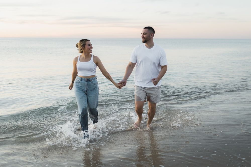 A couple splashes through shallow ocean water at sunset while holding hands and enjoying a romantic beach moment.